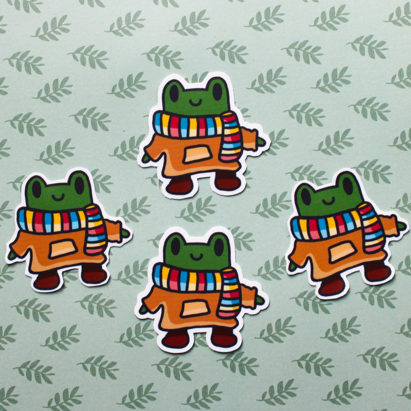 A smiling cartoon frog with a colourful scarf and brown hoodie with brown boots. There are 4 of these stickers placed on a green leaf background.