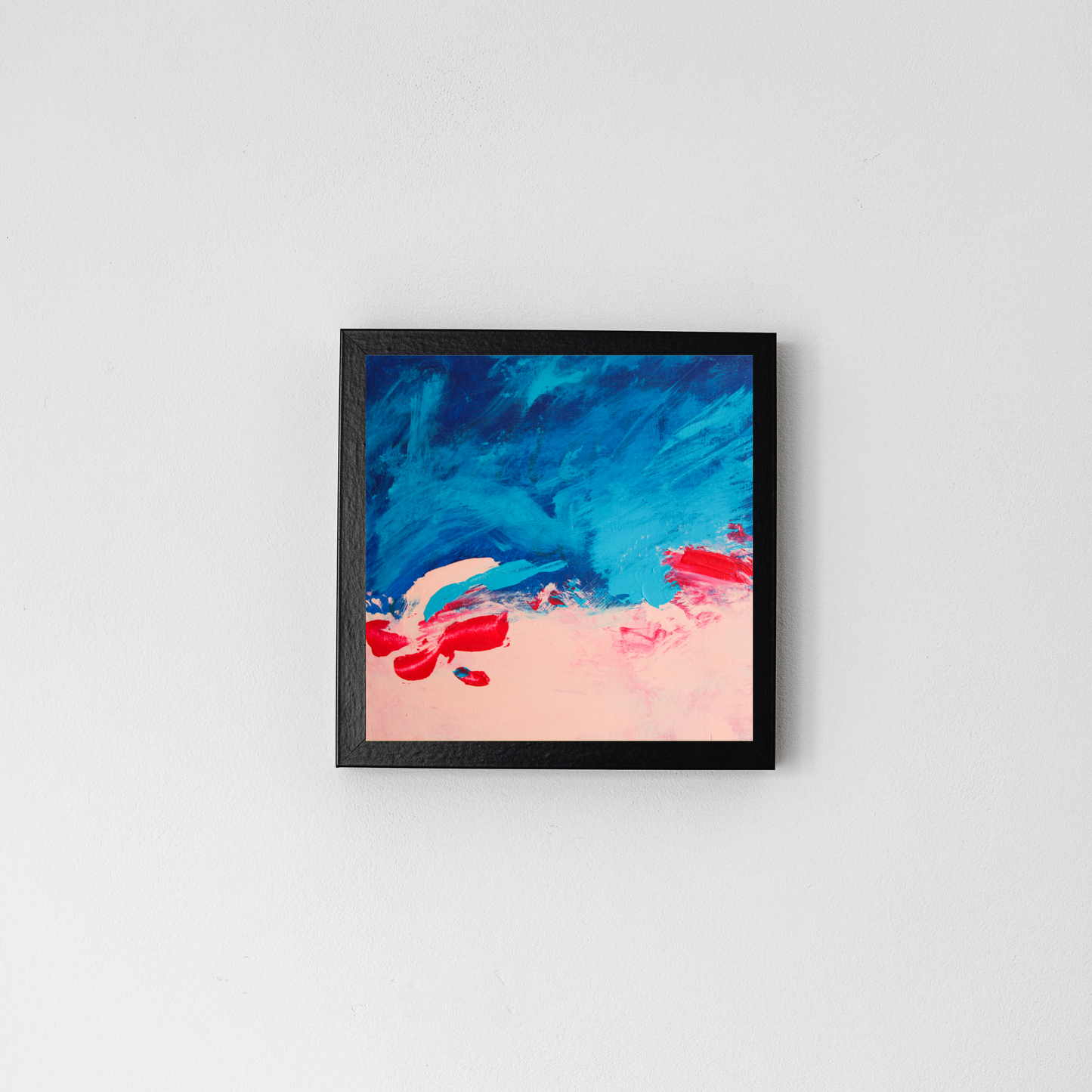 On a grey wall in a black frame is a square blue abstract with pink on the bottom blending into the blue with some red swatches.