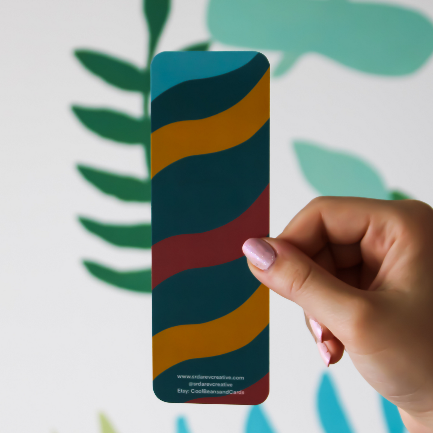The back of the Moosh Jr bookmark. There is "www.srdarevcreative.com @srdarevcreative Etsy: CoolBeansandCards" written on the bottom. The background is a blurred leaf pattern. Has same alternating waves of colour but no mushroom on it.
