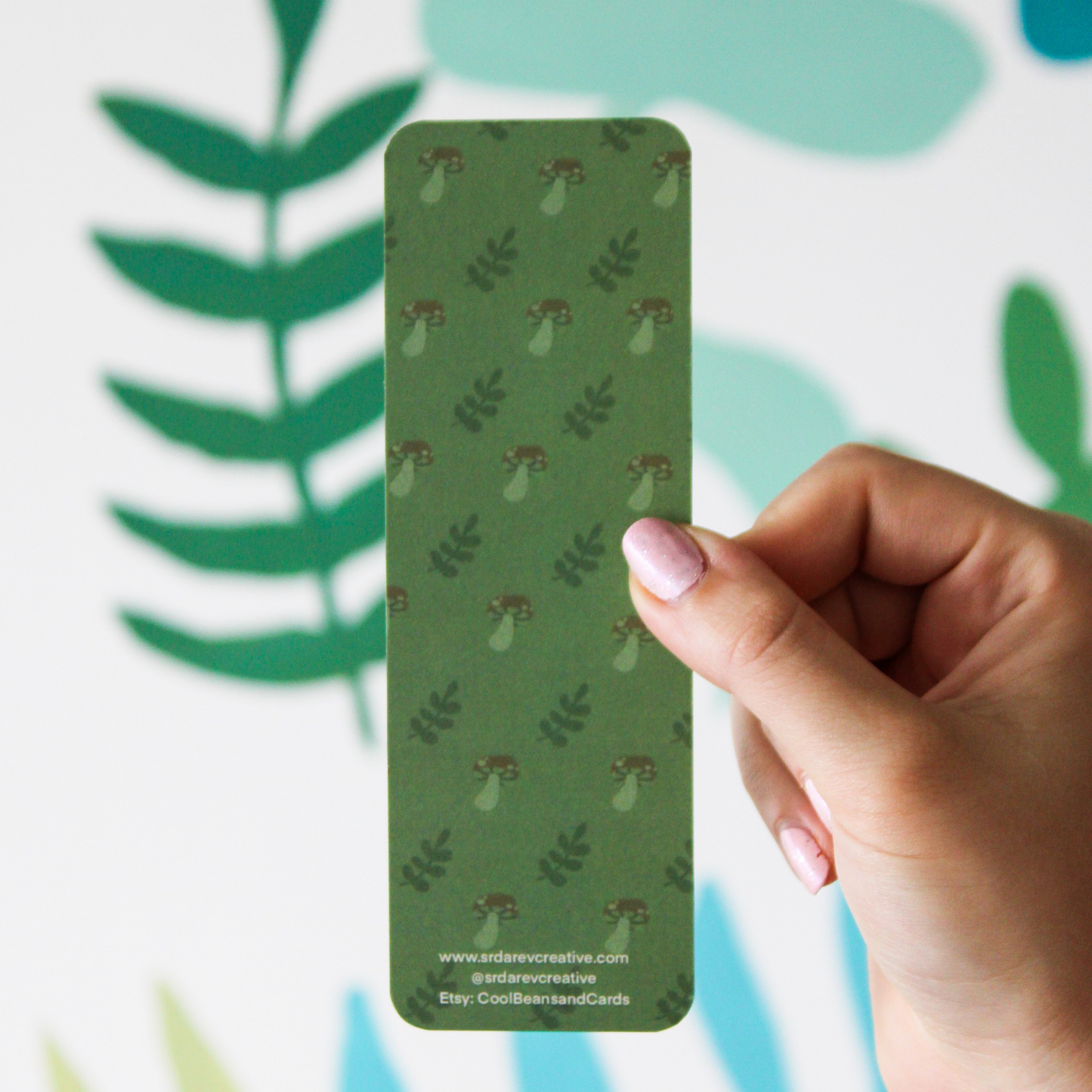 A hand holding the back of the green mushroom bookmark. There is "www.srdarevcreative.com @srdarevcreative Etsy: CoolBeansandCards" written on the bottom. The background is a blurred leaf pattern.