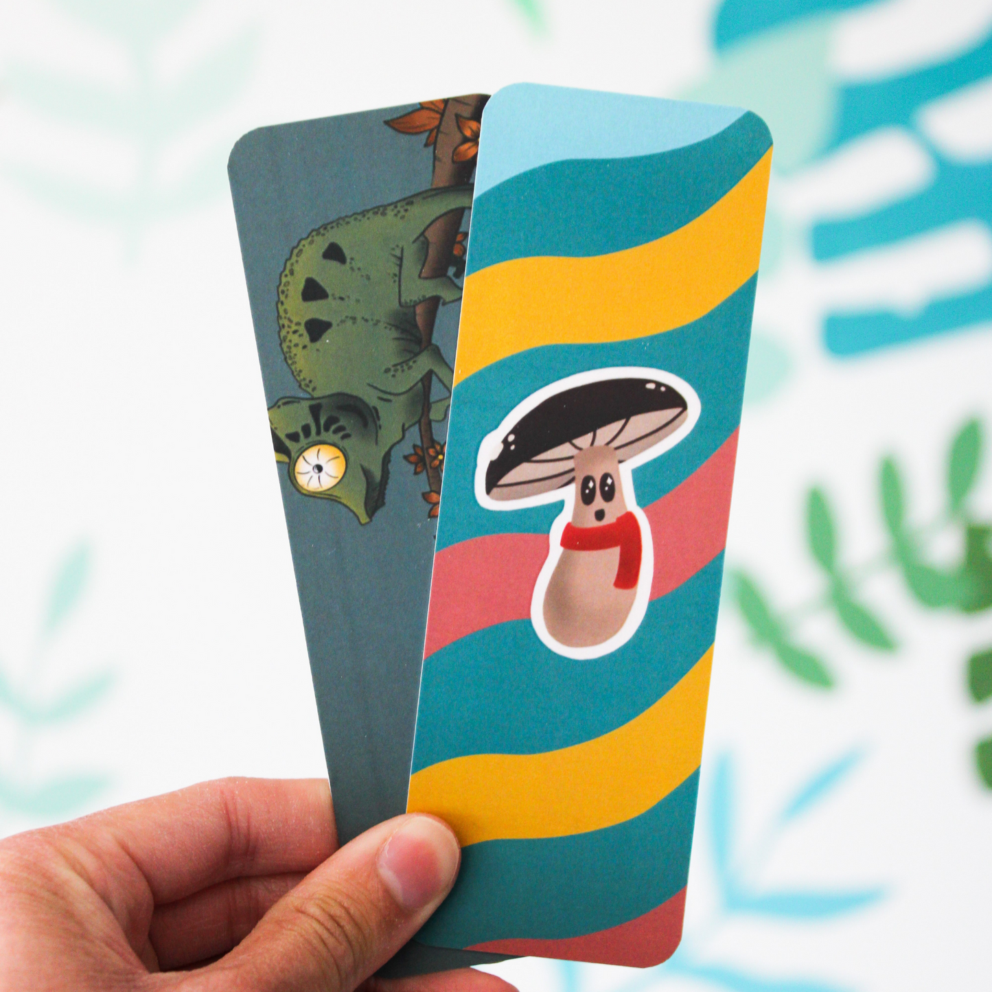 A hand holding two bookmarks. One is the Moosh Jr bookmark, and the other is Charles the Chameleon. Blurred leaf pattern in the background.