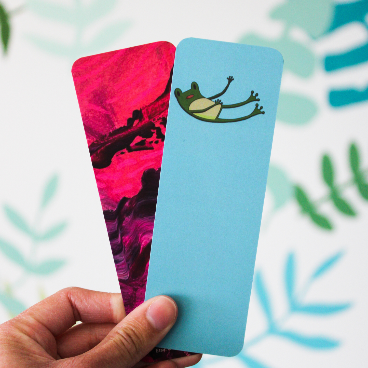 A hand holding the pink and purple bookmark and the Freg Falling bookmark together. The background is a blurred leaf pattern.