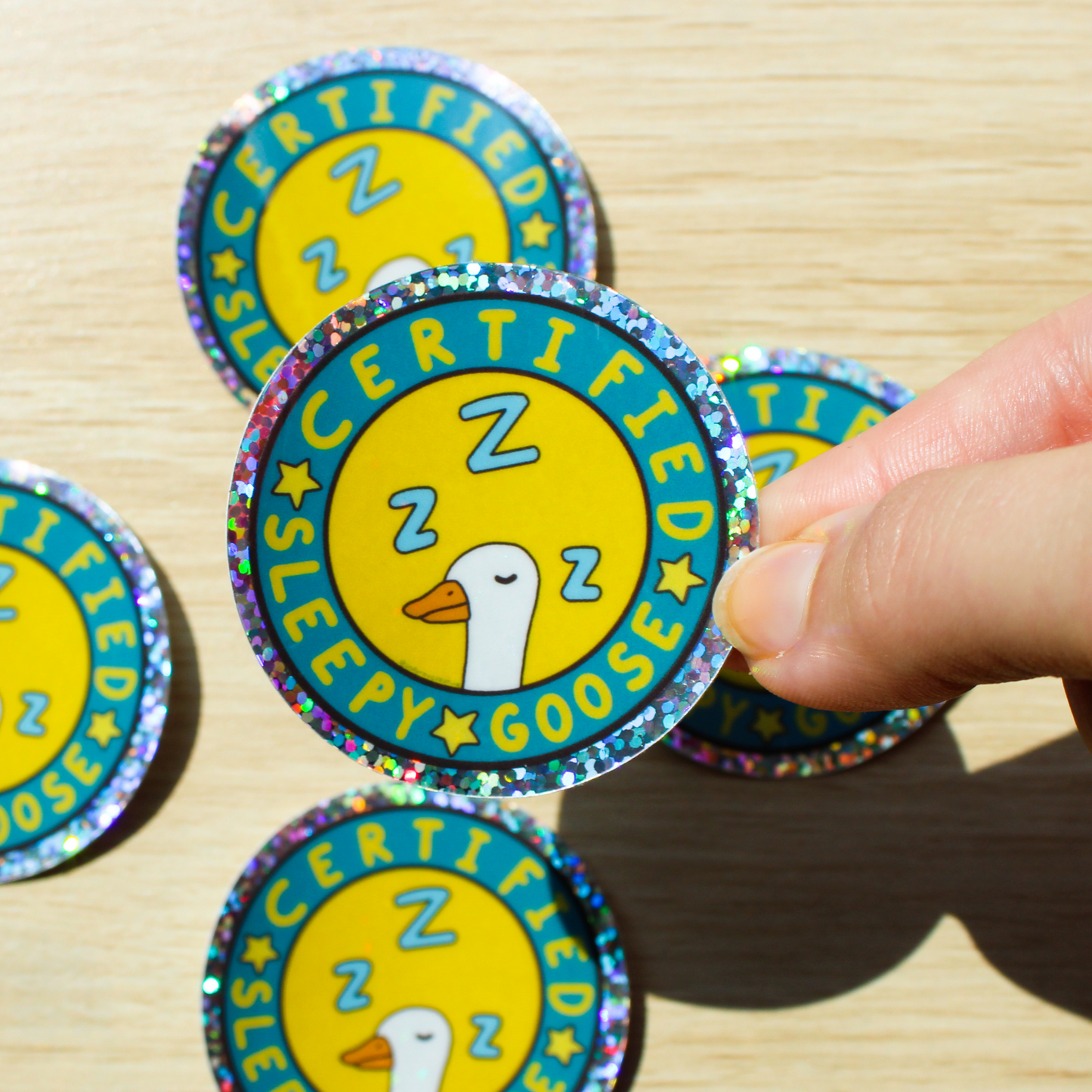 Hand holding a circle holographic sticker that has Certified Sleepy Goose written in a circle with stars and a white goose with Z's sleeping on a yellow background. Background is more certified sleepy goose stickers on a wooden table.