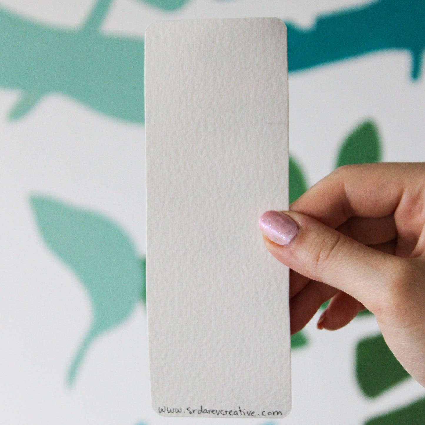 A blurred leaf pattern background. In the front is a hand holding a white bookmark with the words "www.srdarevcreative.com" written in black on the bottom.