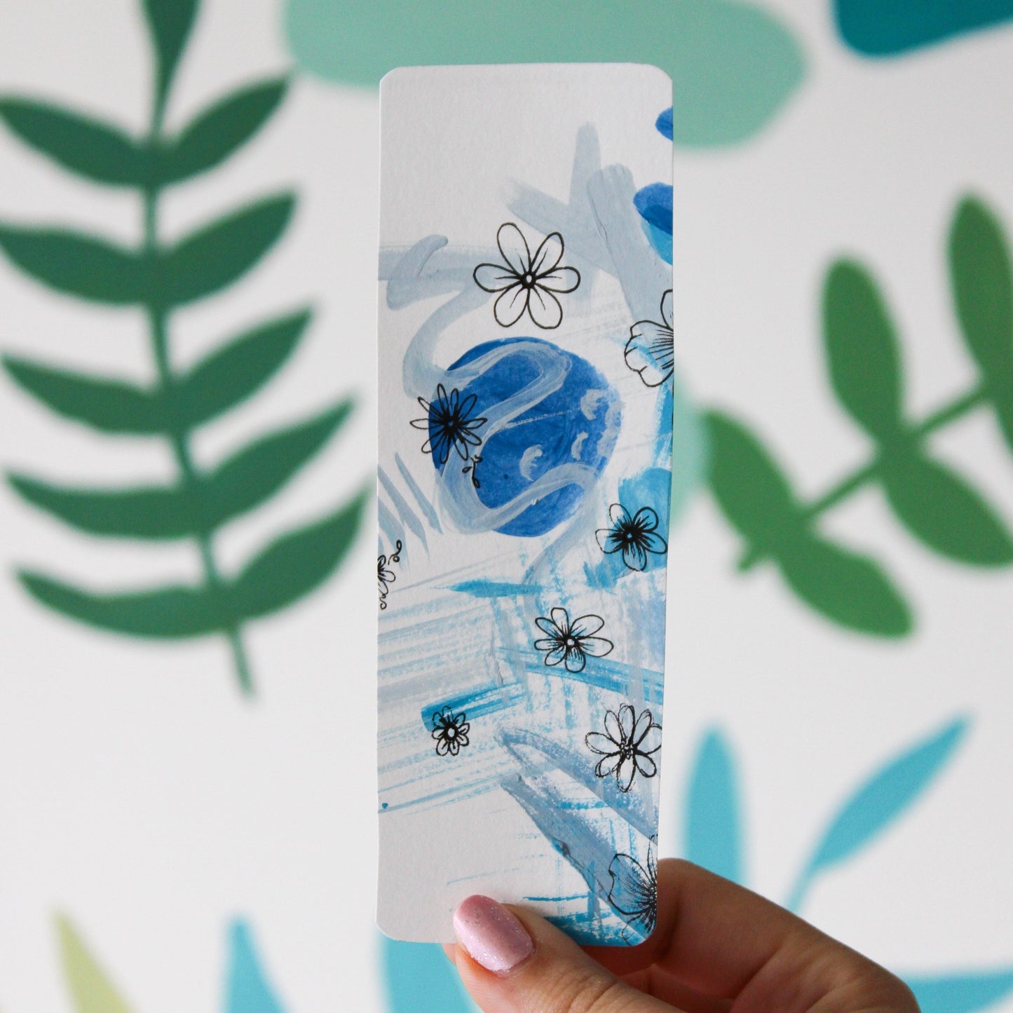A blurred leaf background. In front is a hand holding a white bookmark with blue abstract paintings and flowers drawn in black ink.