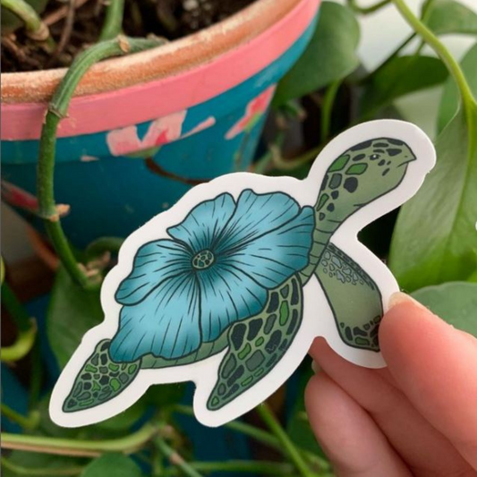 A leafy plant in the background. In the front is a hand holding a turtle sticker with a blue flower on it in place of a shell.