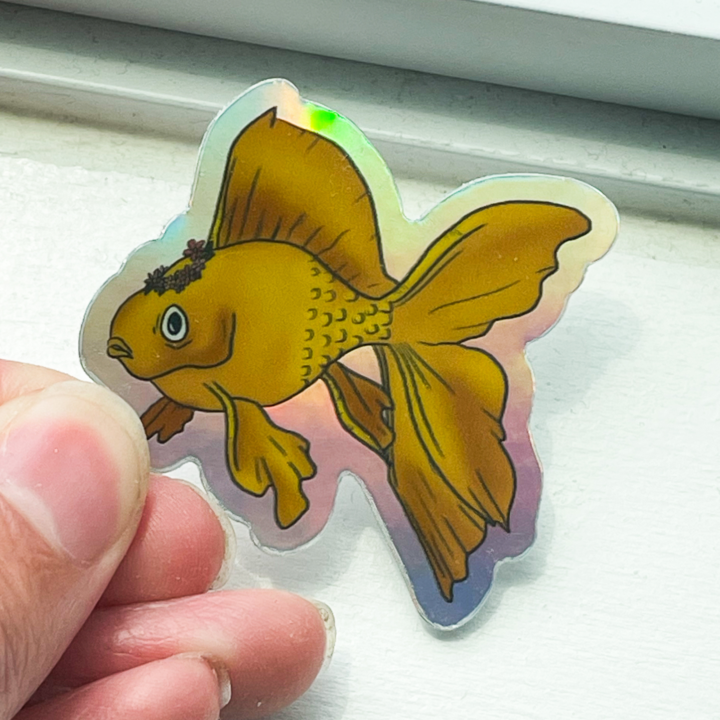 A white window sill background. In the front is a hand holding a holographic sticker that has an orange goldfish wearing a pink flower crown.