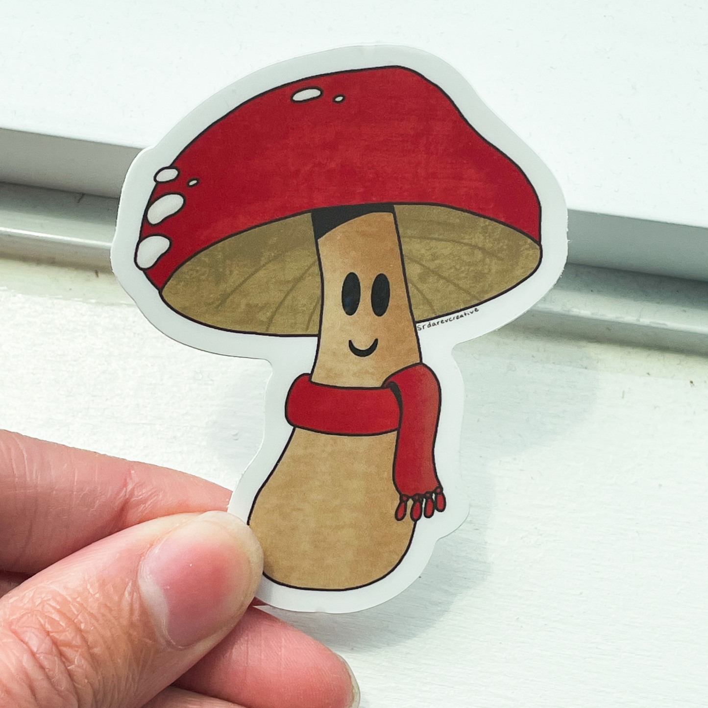 A white window sill background. In the front is a hand holding a mushroom sticker that has a red cap and matching red scarf with a smile on its face.