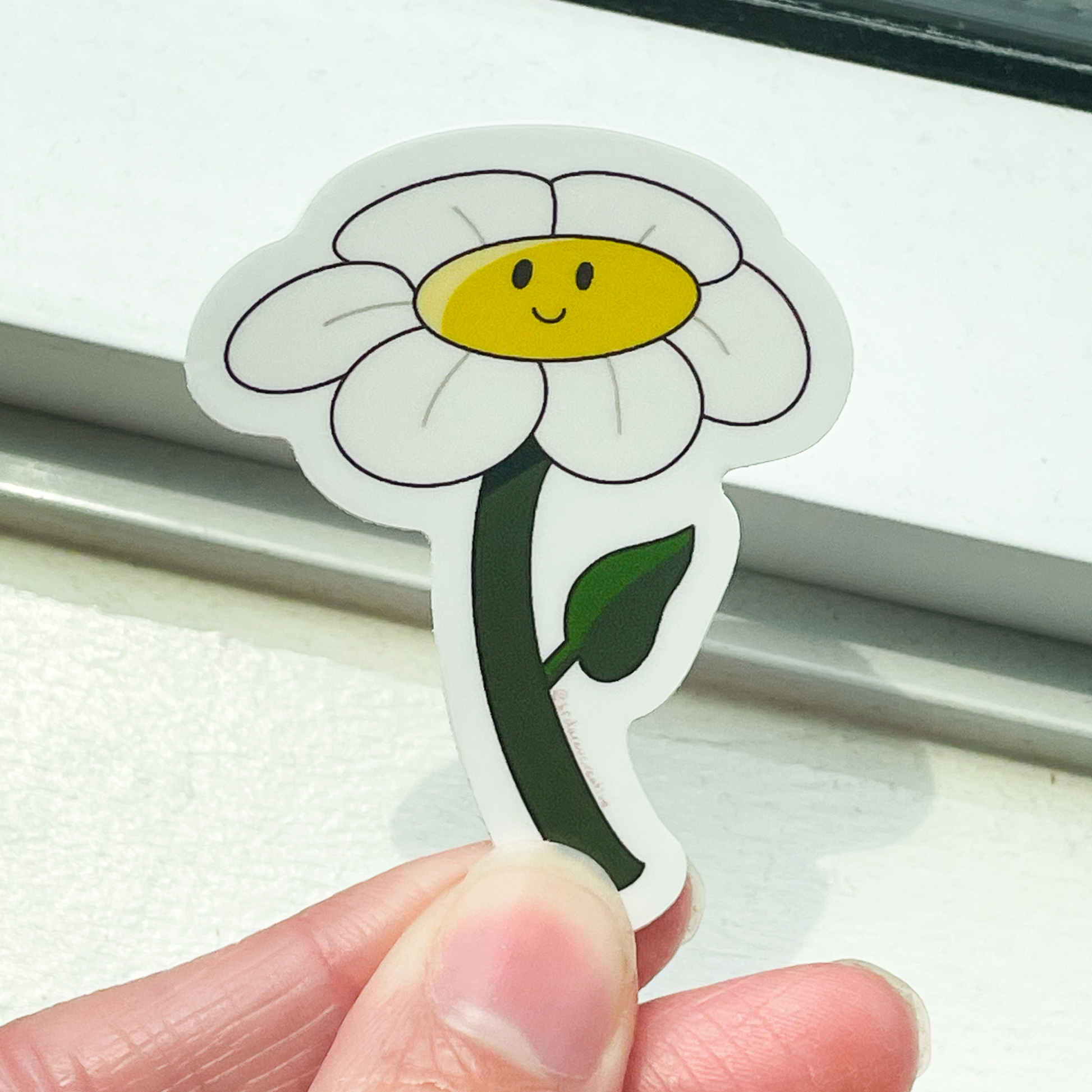 A white window sill background with a flower sticker on top that has white petals, a dark green stem, and a yellow centre with a smiling face.