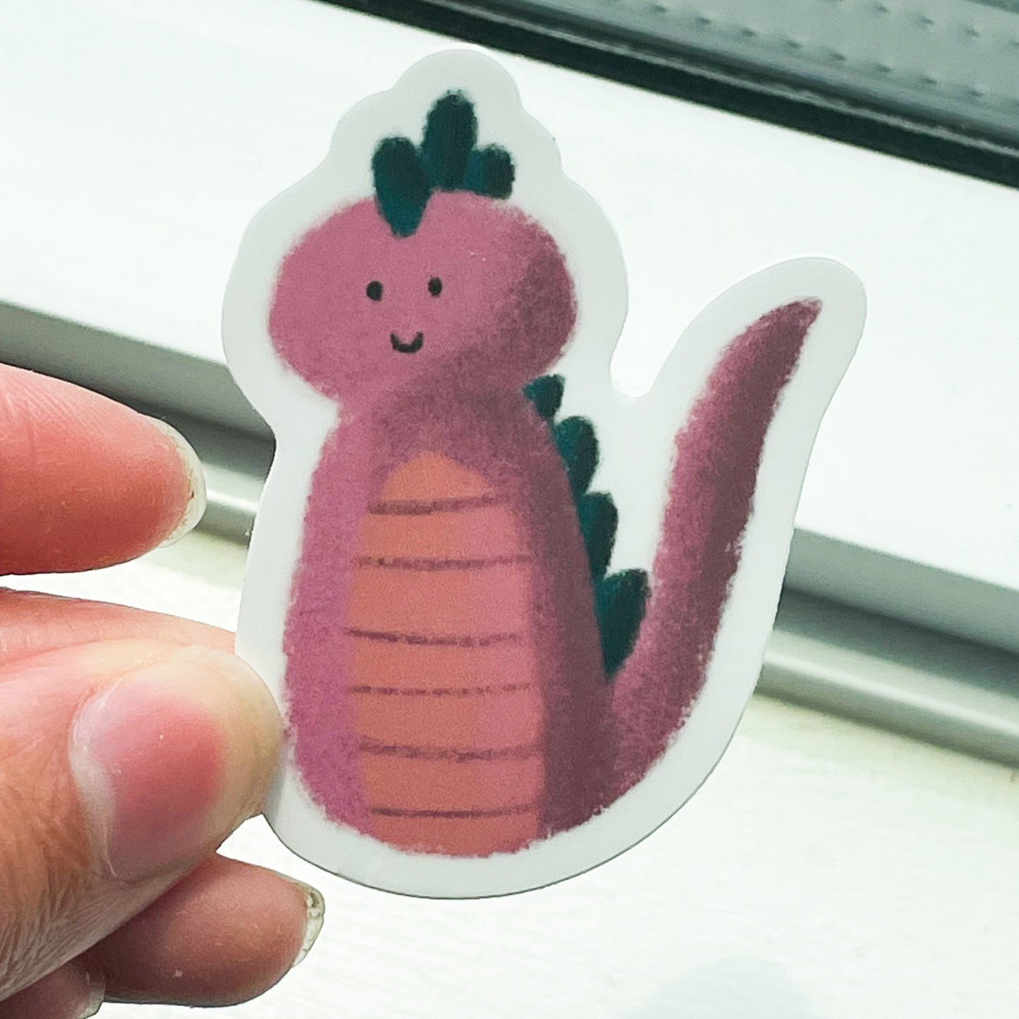 A hand holding a sticker that has a smiling pink monster on it with dark green spikes and a long pink tail. The background is the bottom of a white window sill.