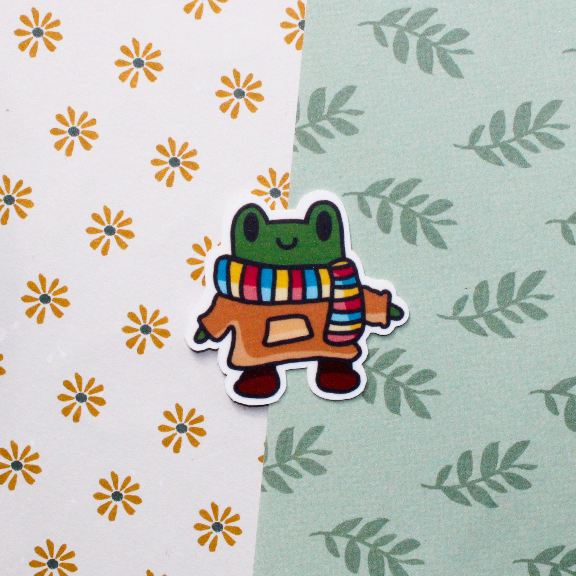 A smiling cartoon frog with a colourful scarf and brown hoodie with brown boots. Background is a flower and leaf pattern.