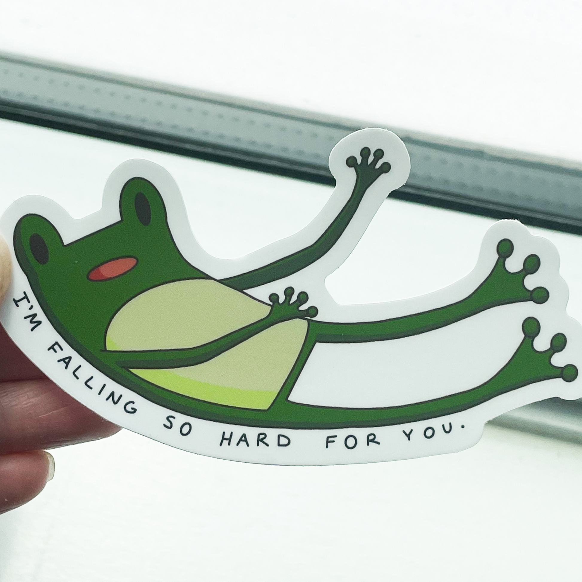 A hand holding a sticker of a green cartoon frog falling with the words "I'M FALLING SO HARD FOR YOU." underneath on a white background.