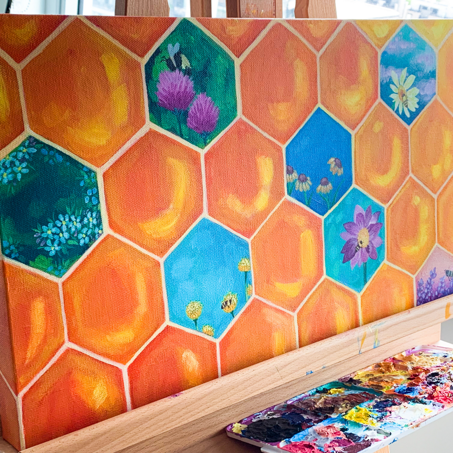 An easel with an orange honeycomb painting wiht some honeycombs having different colours and flowers and bees painted in them. Under the easel is a paint palette.