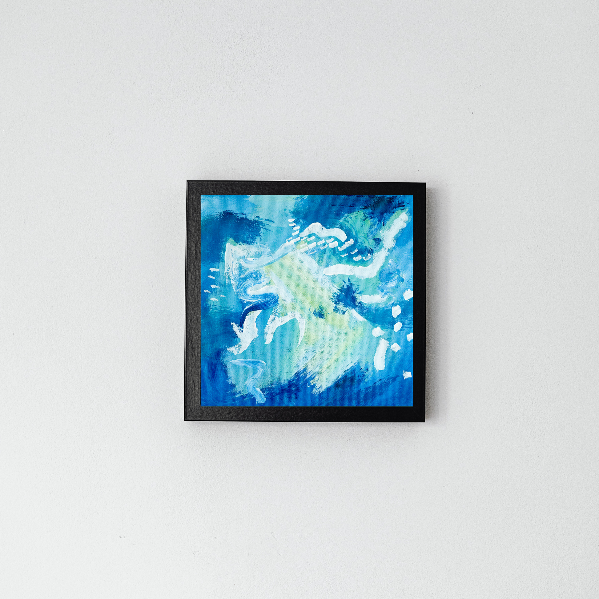 An abstract blue painting is pictured in a black frame on a grey wall with accents of white and yellow in a black frame on a grey wall