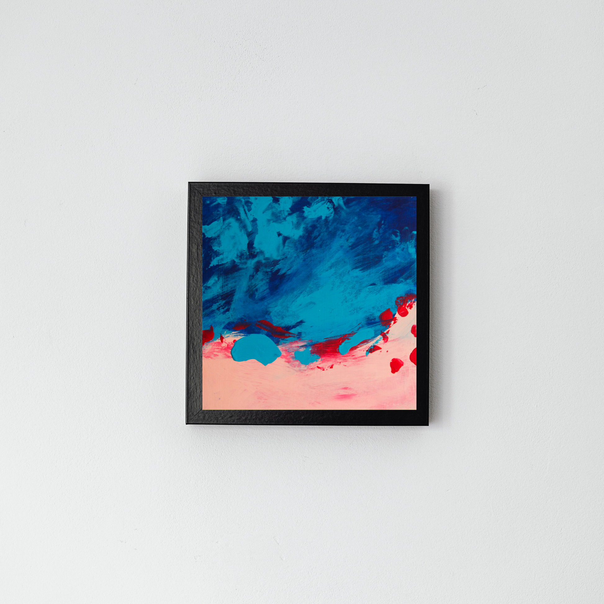 On a grey wall in a black frame is a square blue abstract with pink on the bottom blending into the blue with some red swatches.