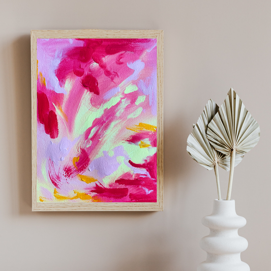 A tan wall with a white vase and paper leaves on the right. On the left is a wooden frame with a bright pink, purple, and yellow abstract painting in it.