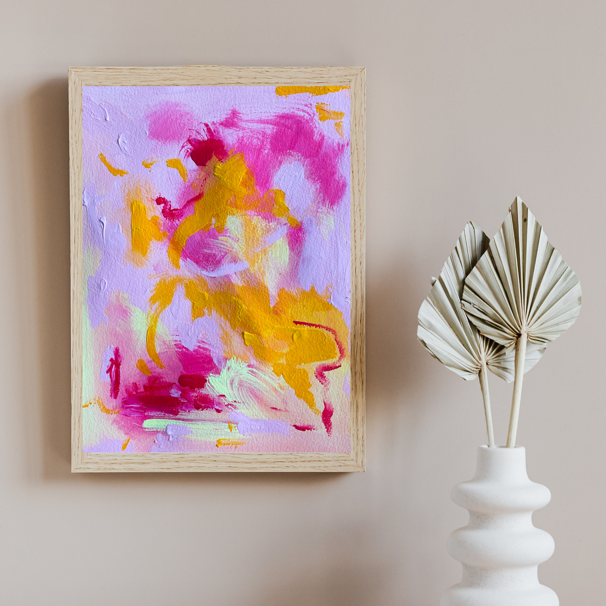 A tan wall with a white vase and paper leaves on the right. On the left is a wooden frame with a bright pink, purple, and orange abstract painting in it.