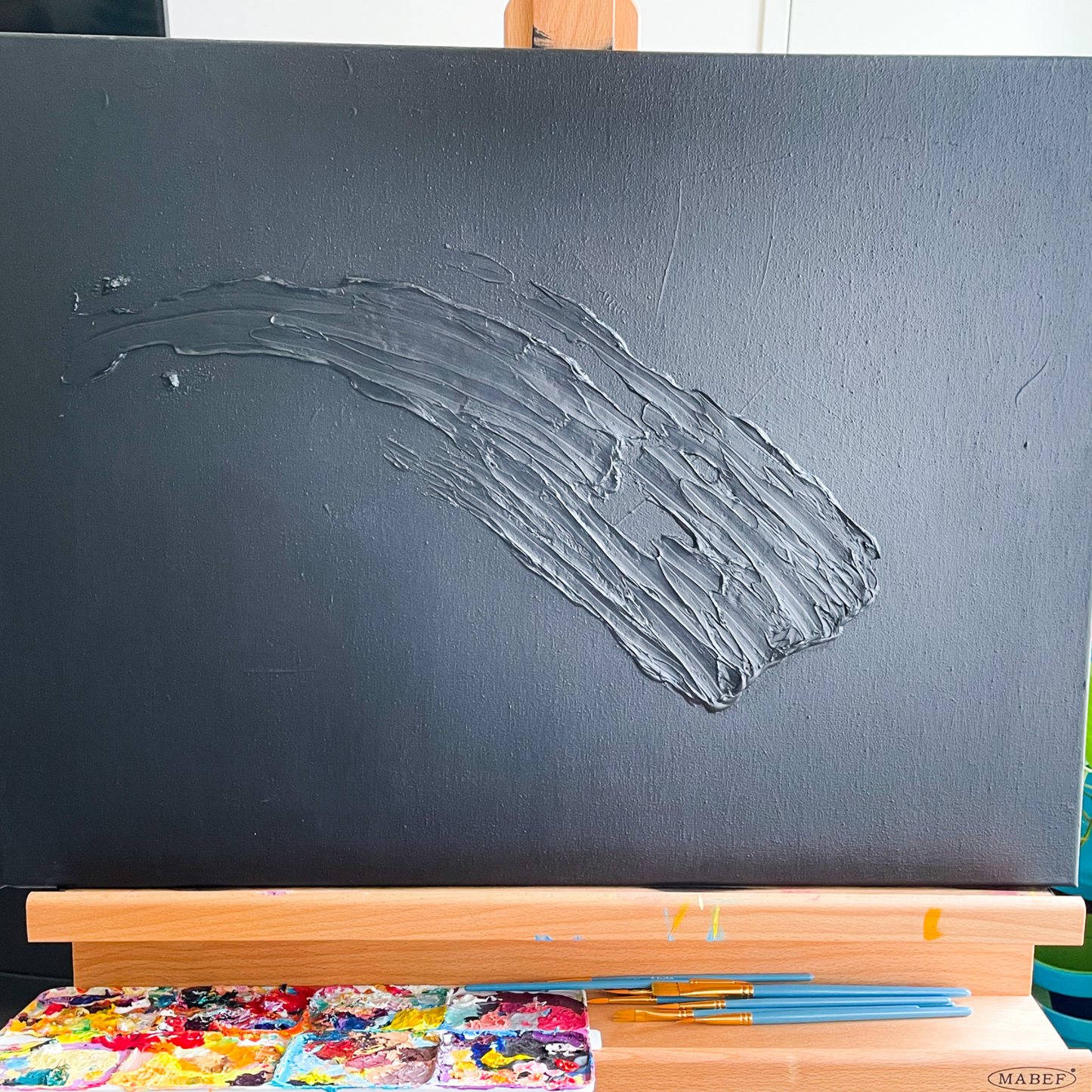 On an easel is a black painting with a large textured downward swoosh. Underneath the easel is a paint palette and paint brushes.