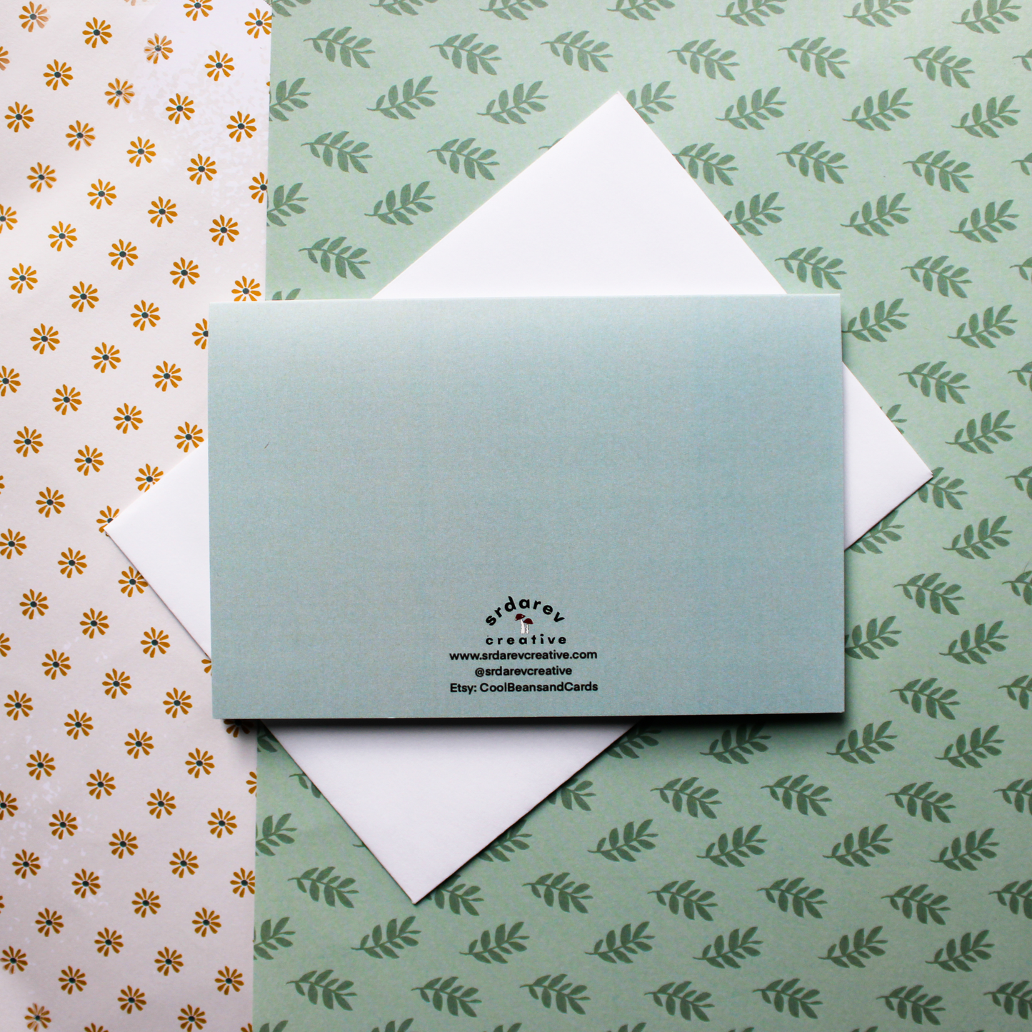 A light blue card on top of a white envelope with the "Srdarev Creative" logo on the back with mushrooms in the middle. "www.srdarevcreative.com" "@srdarevcreative". The background is a green leaf pattern and yellow floral pattern. 