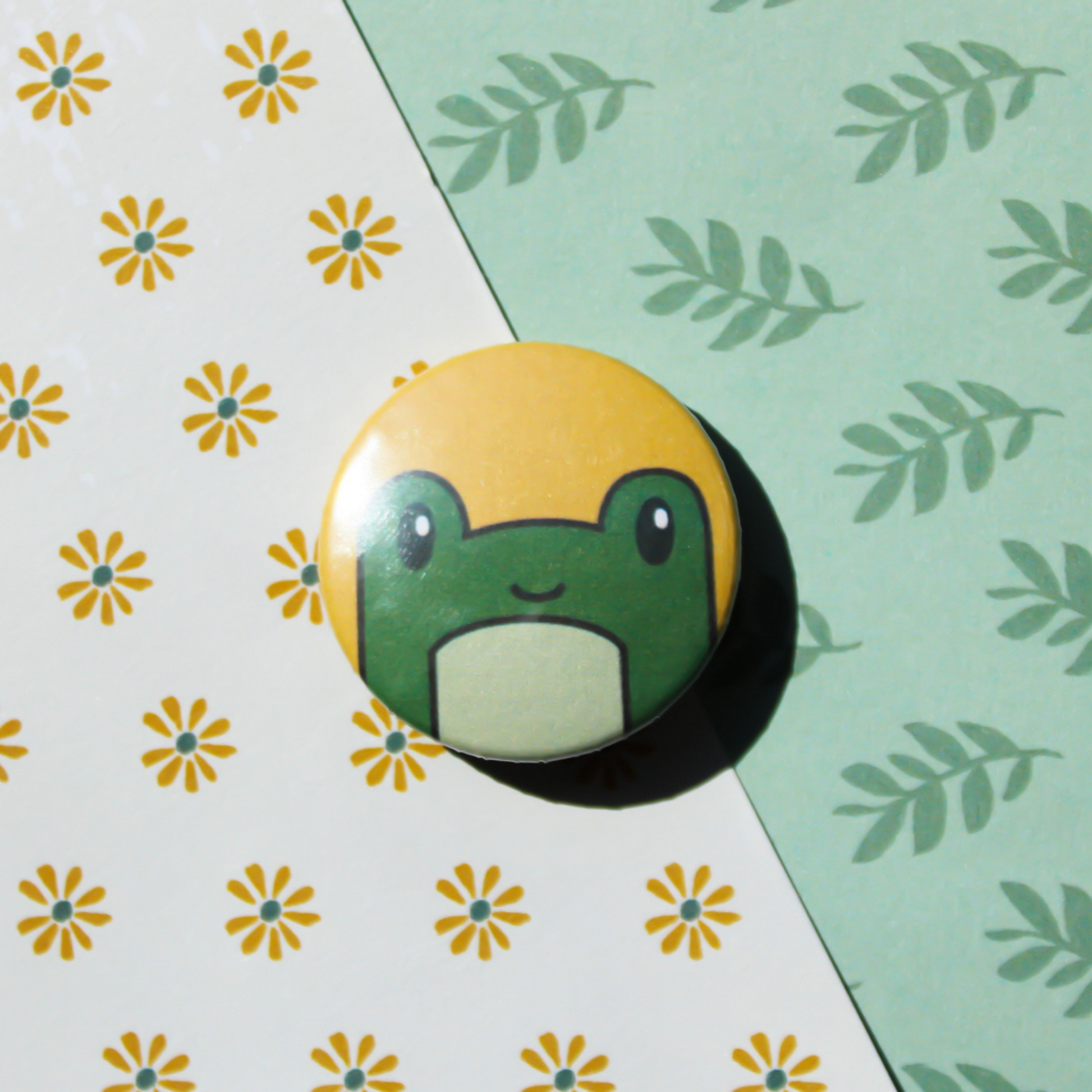 A leaf and flower pattern background. On top is a yellow circle pin with a green cartoon frog's head smiling.