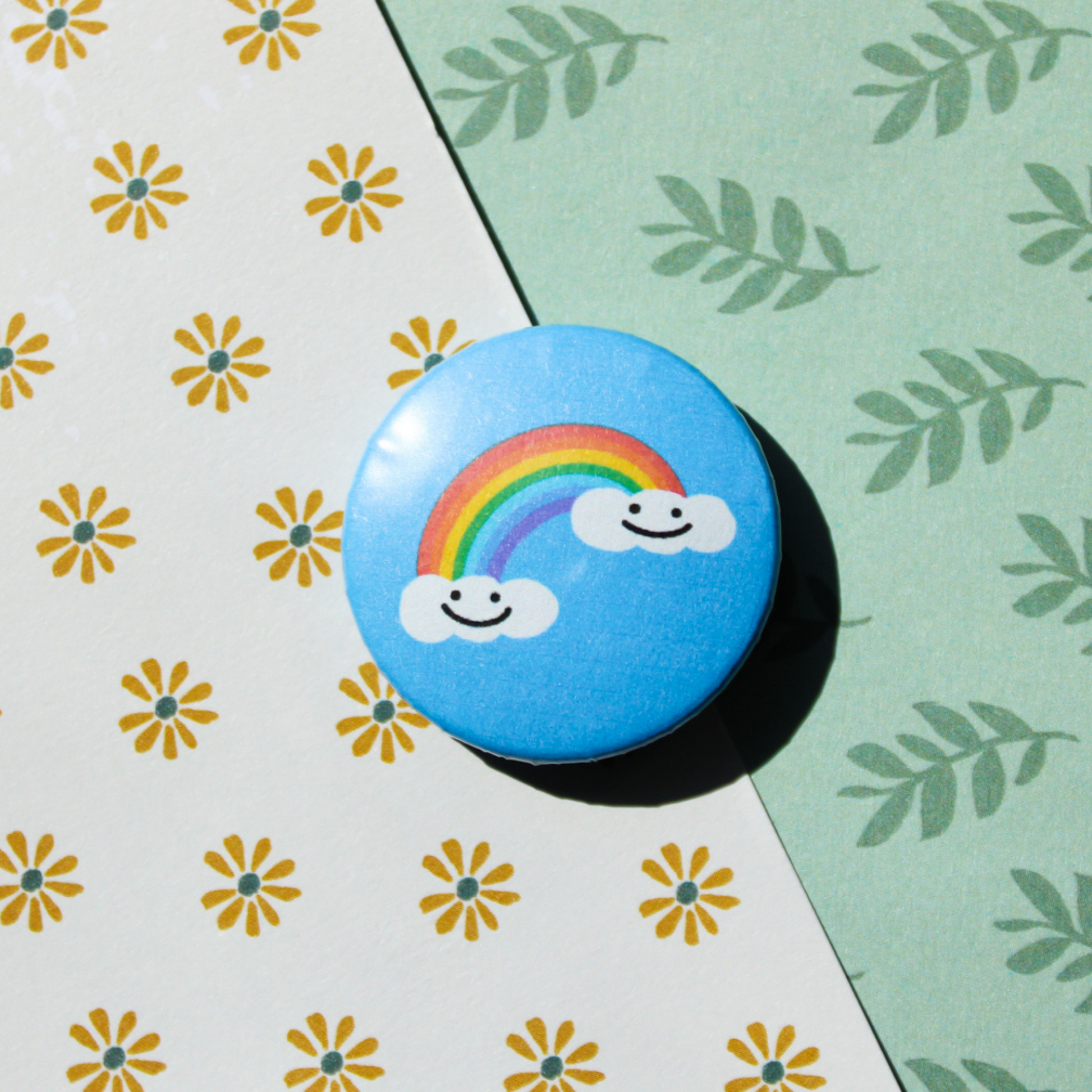 A leaf and floral pattern background. On top is a blue circle button with two smiling white clouds. In between those clouds is a bright rainbow.