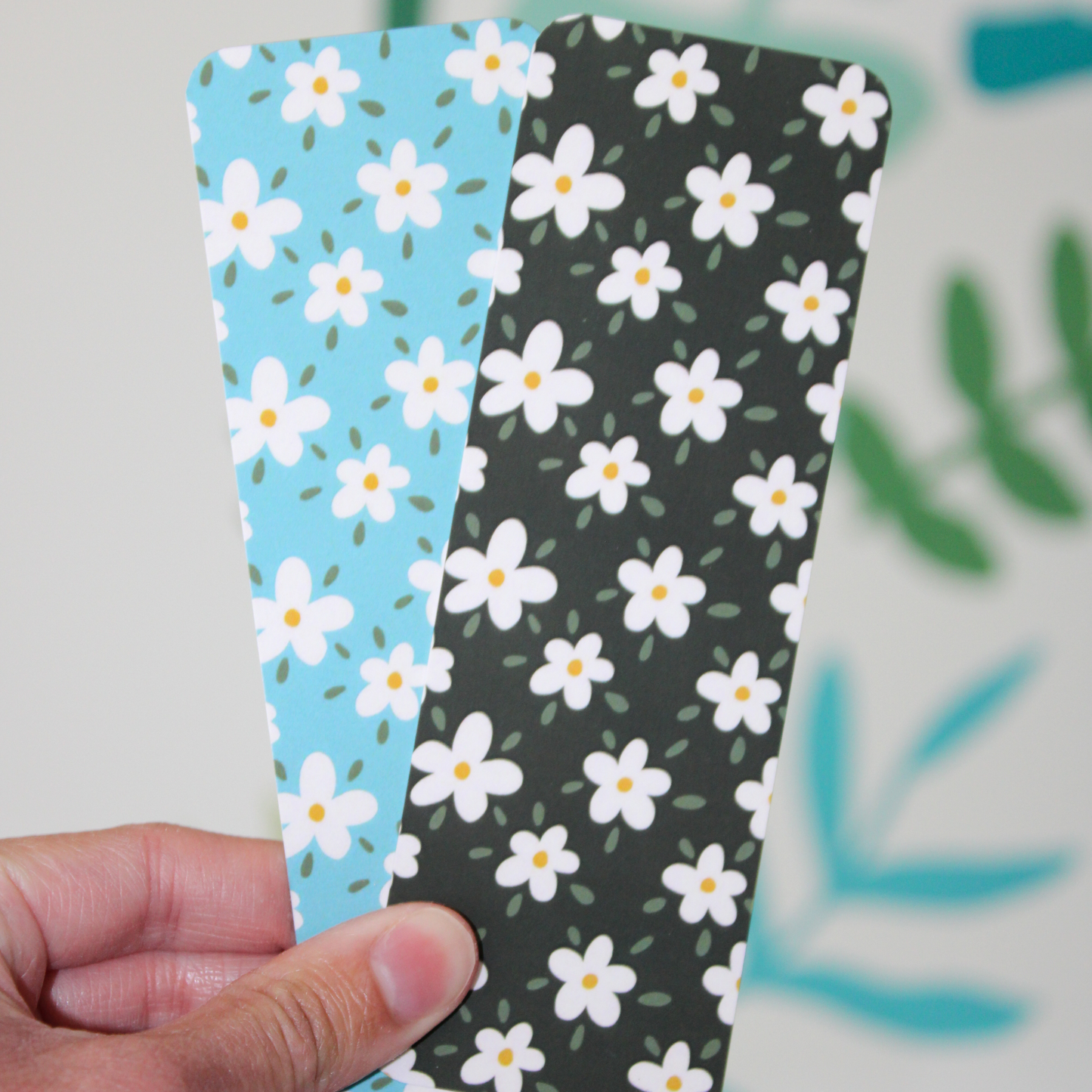 A hand holding two bookmarks, one being the blue daisy pattern and one being the black bookmark with a daisy pattern. The background of the photo has blurred leaf patterns.
