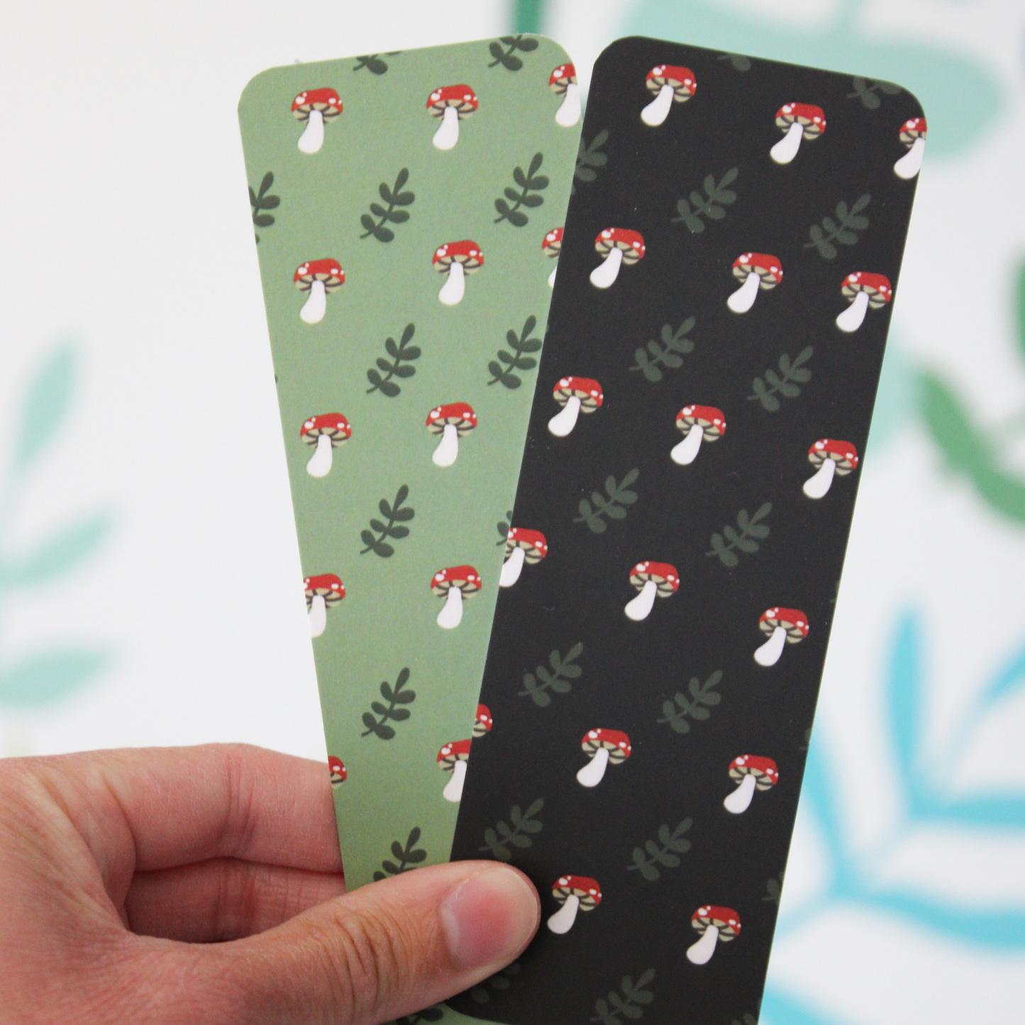 A hand holding two bookmarks. One has the green mushroom pattern and one has the black mushroom pattern. 