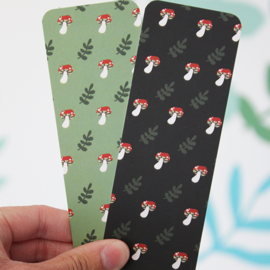 Hand holding two bookmarks, one is the green mushroom bookmark and one is the black mushroom bookmark.