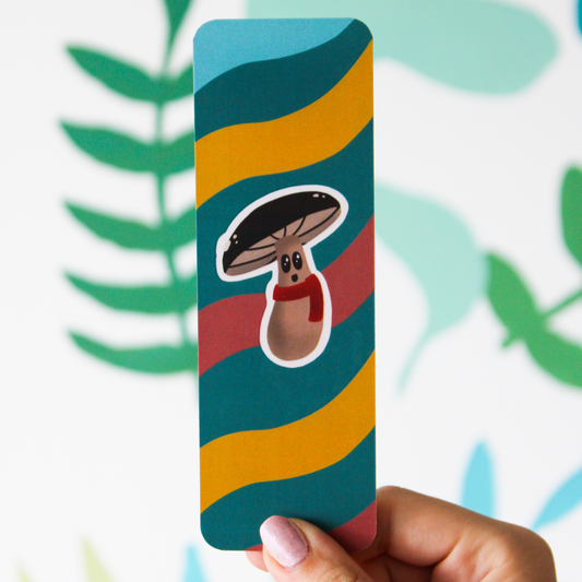 Hand holding a bookmark. Bookmark has alternating light blue, dark blue, yellow, and coral waves. In the middle is a happy brown mushroom wearing a red scarf with a white outline.