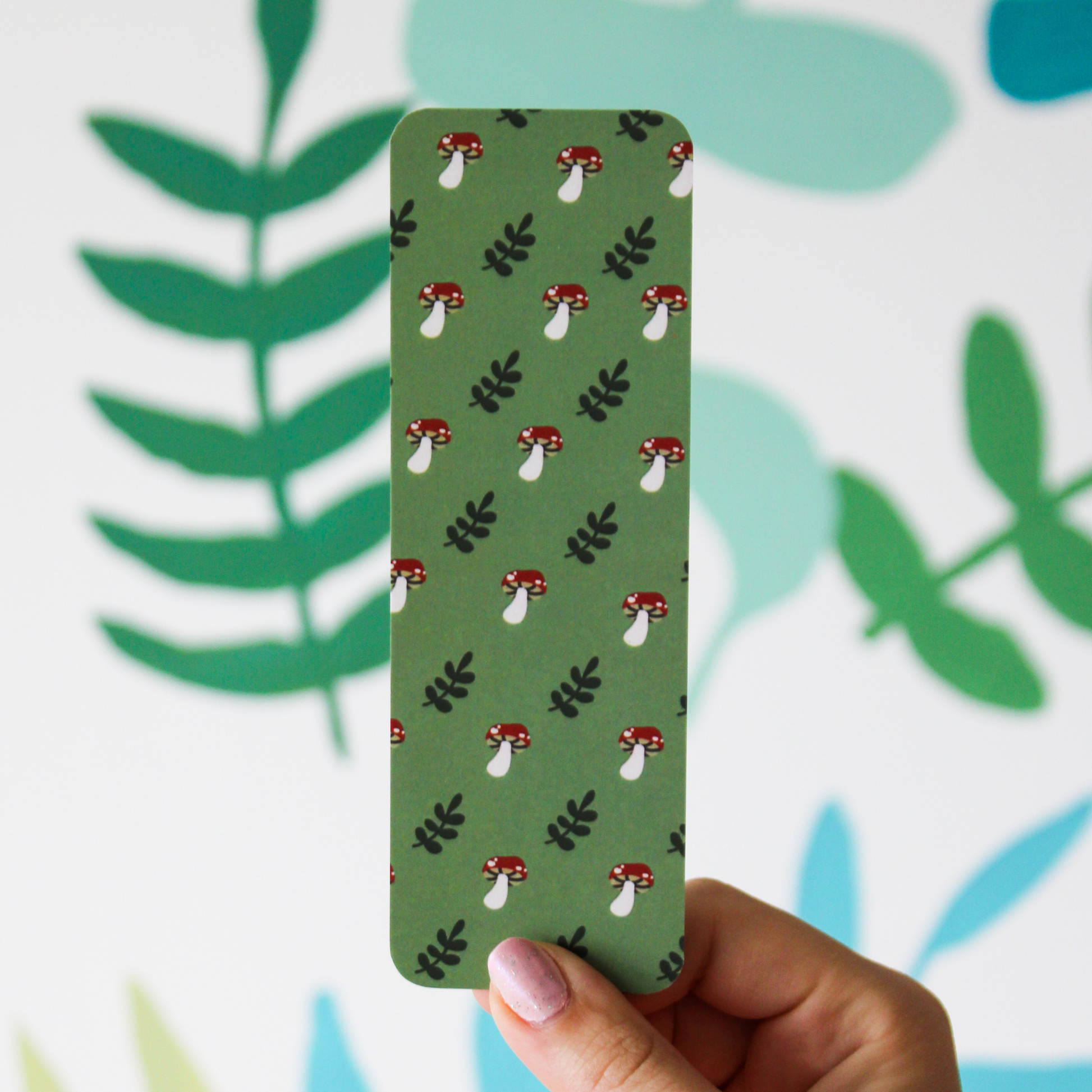 A hand holding a green bookmark with an alternating mushroom and leaf pattern. The background has a blurred leaf pattern.