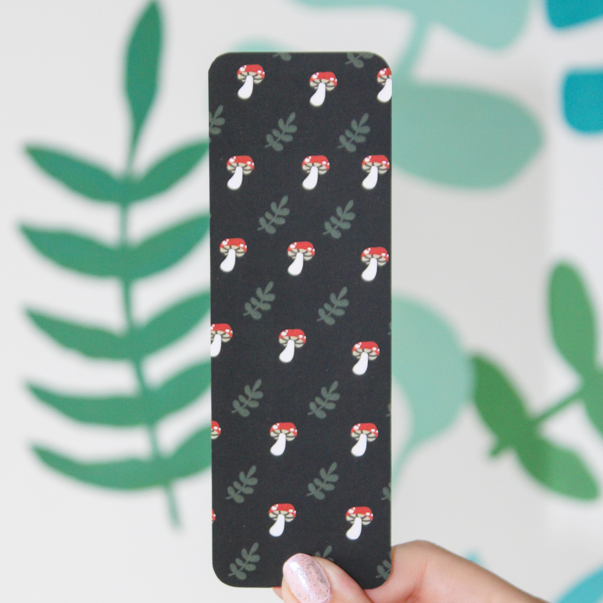 Hand holding black bookmark with a pattern of red mushrooms and leaves on it. Blurred leaf pattern in the background.