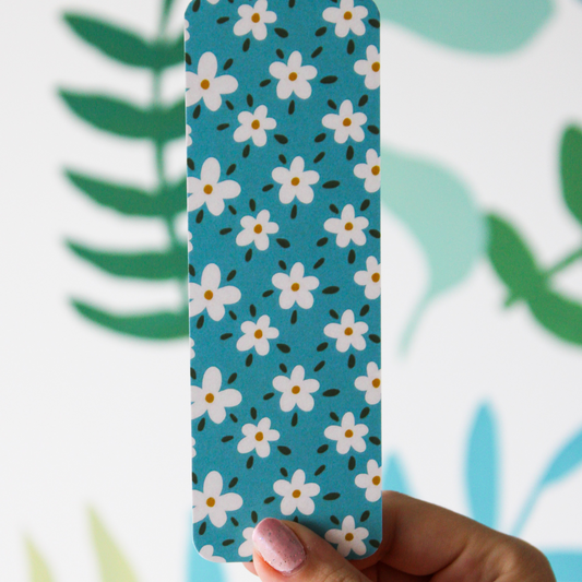 A hand holding a blue bookmark with daisies and dark green leaf pattern. The background of the photo has blurred leaves.
