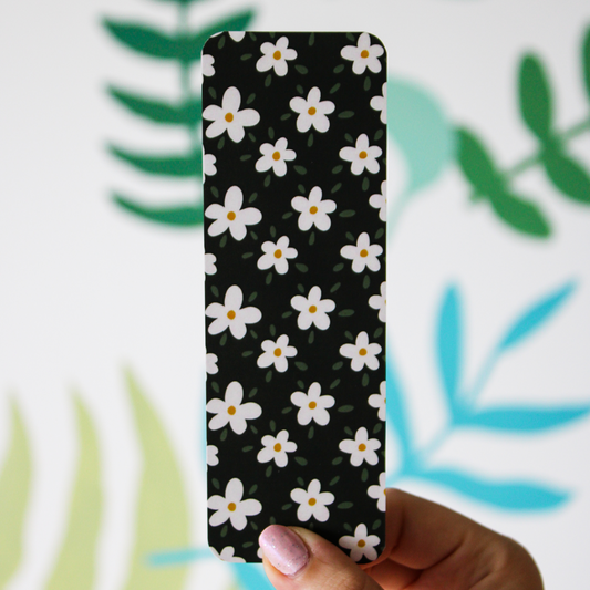 A hand holding a black bookmark with white daisies and little green leaves throughout. Blurred leaf pattern is in the background.