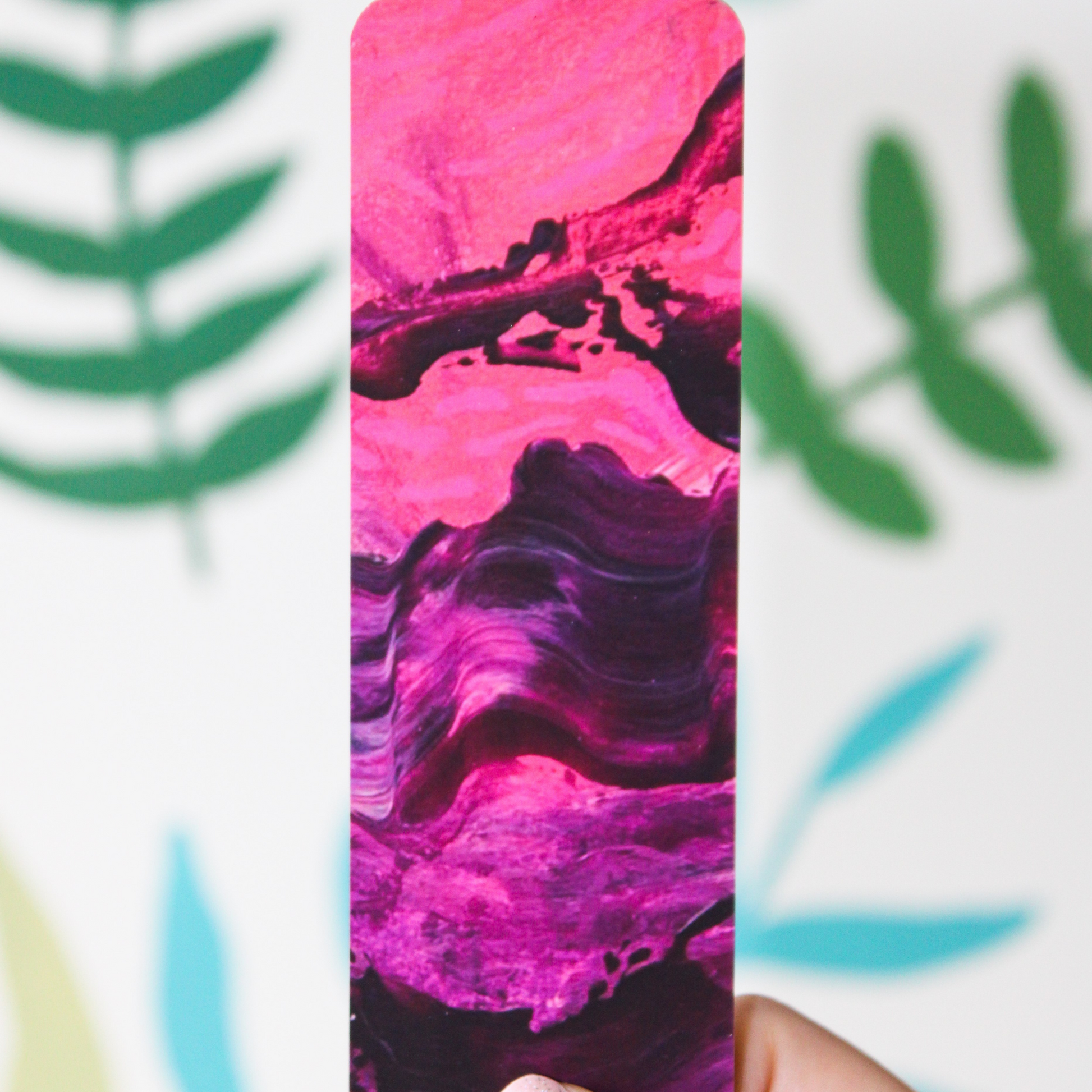 A picture of a pink and purple abstract bookmark with bright pink and the top turning into purple on the bottom. The background is a blurred leaf pattern.