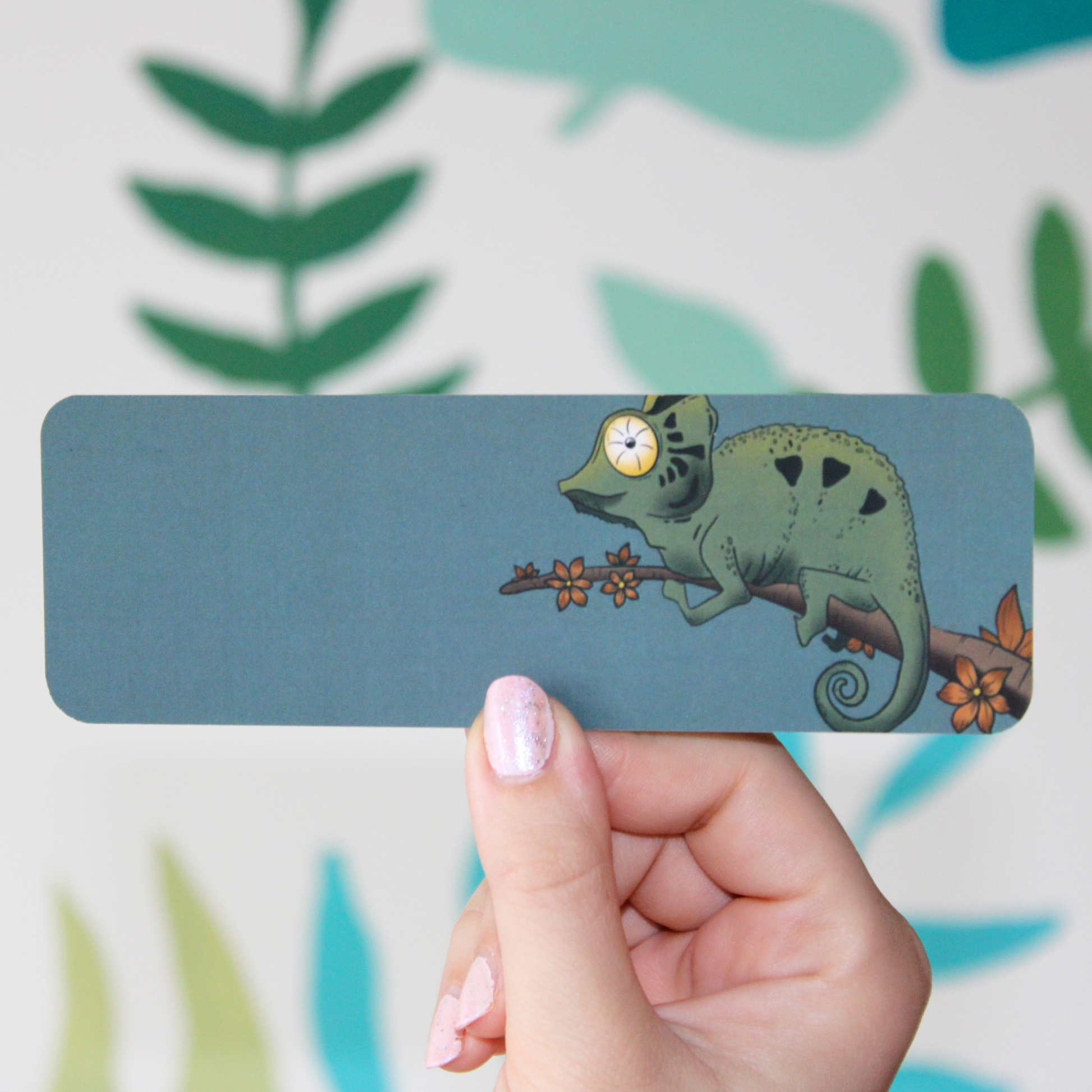 A hand holding a blue/grey bookmark with a green chameleon on a branch with orange flowers. There's a blurred leaf pattern in the background.
