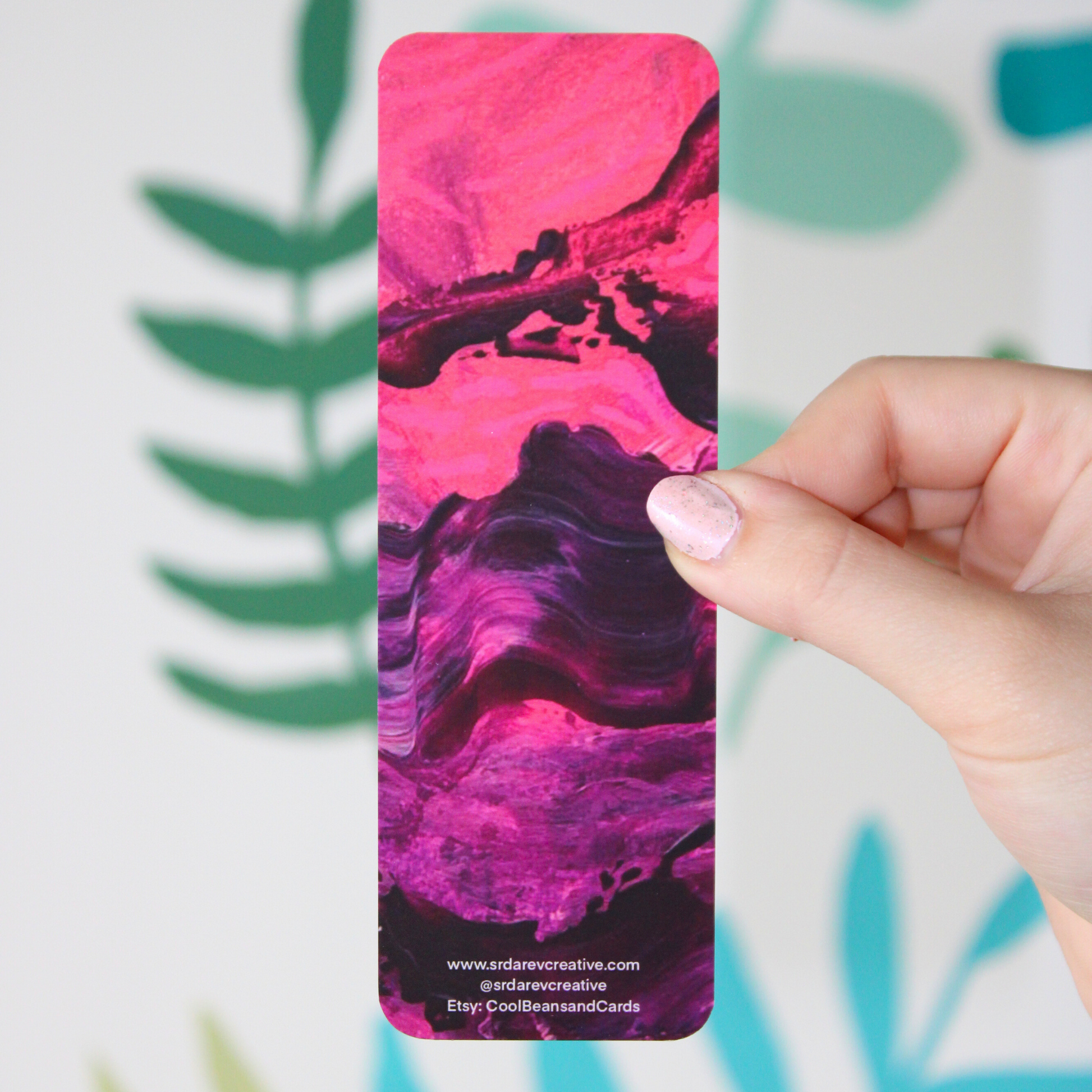 A picture of the back of a pink and purple abstract bookmark with bright pink and the top turning into purple on the bottom. There is "www.srdarevcreative.com @srdarevcreative Etsy: CoolBeansandCards" written on the bottom. The background is a blurred leaf pattern.