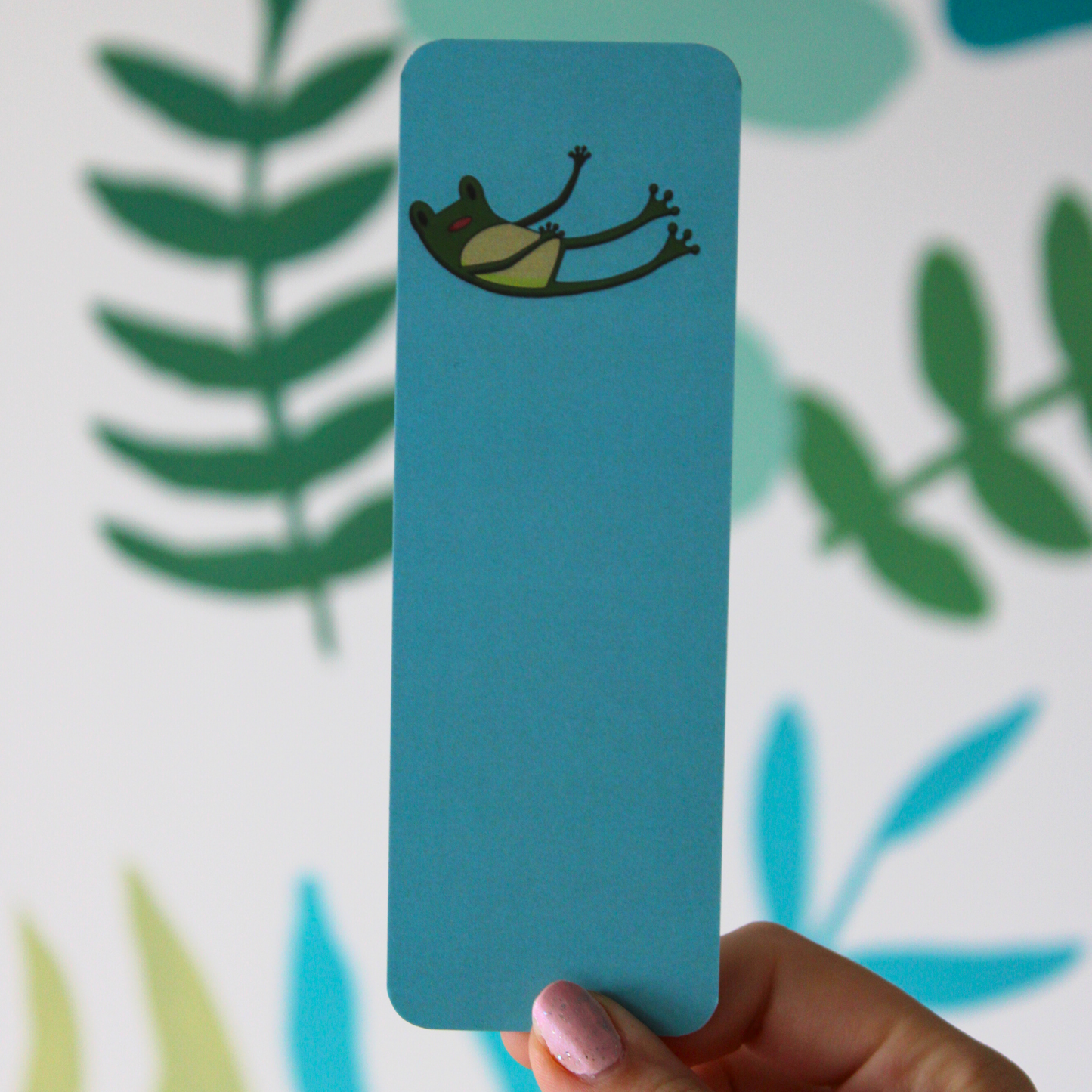 A hand holding a blue bookmark with a cartoon frog falling. The background is a blurred leaf pattern.