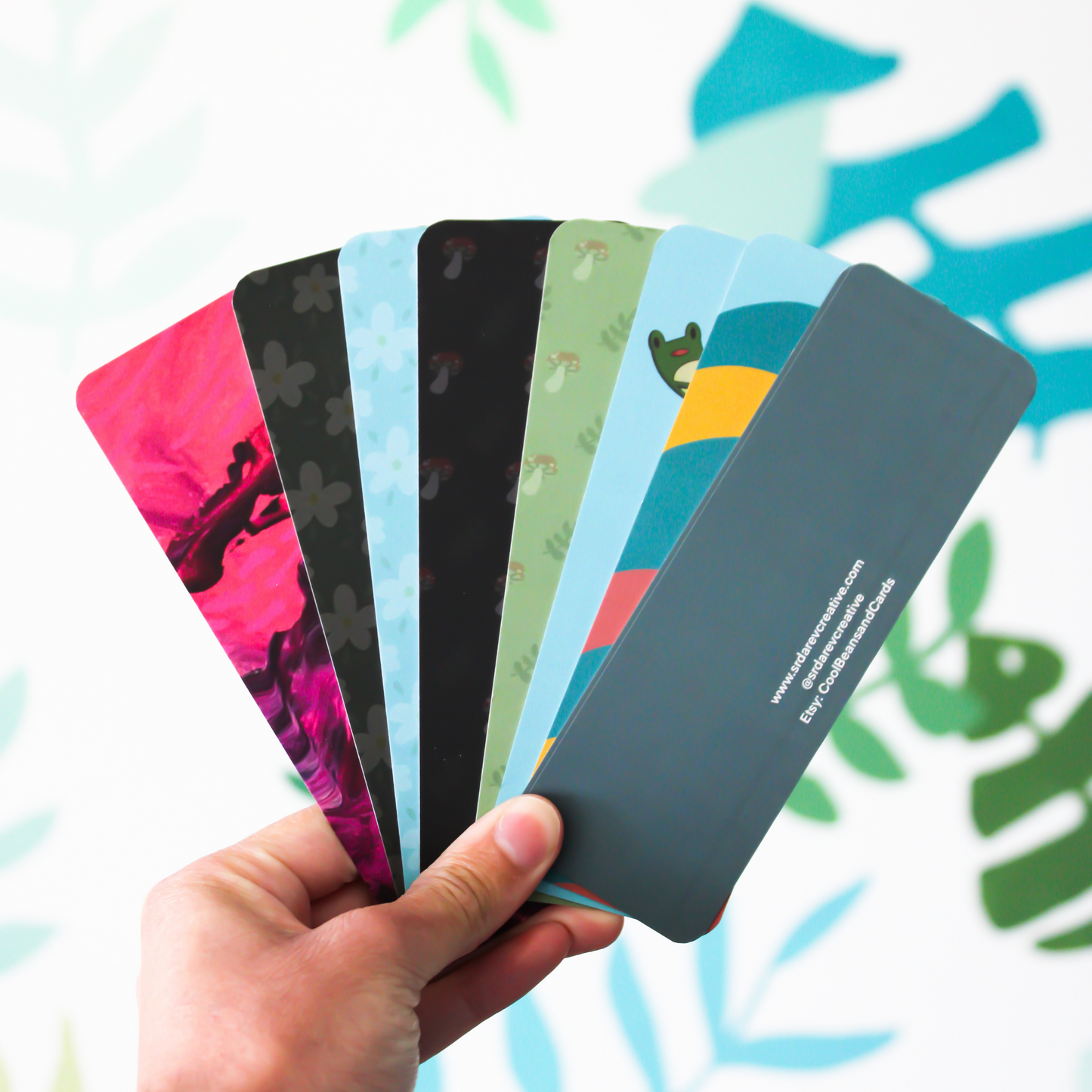 A hand holding the backside of 8 different bookmarks in a fan formation. The background is a blurred leaf pattern.
