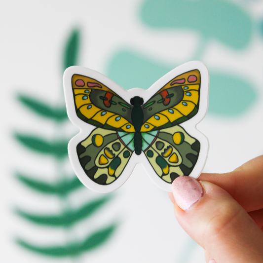 A butterfly sticker with blue, yellow, and green highlights is seen held in a hand on a blurred green leaf background