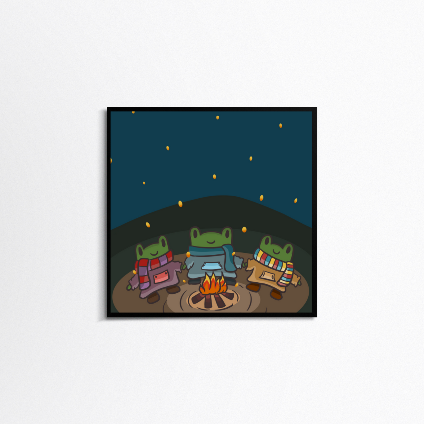 White background with a square black frame. In the frame is a print with a dark blue sky and green and brown forefront. There are three cartoon frogs smiling holding hands in sweaters and scarves around a fire.