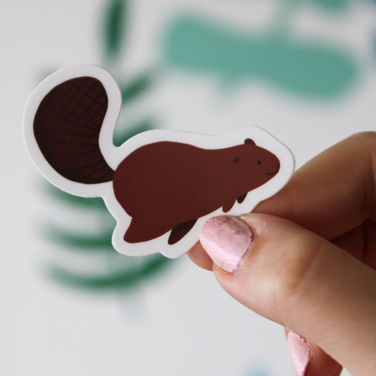 A blurred leaf background. A hand is a holding a smiling beaver sticker.