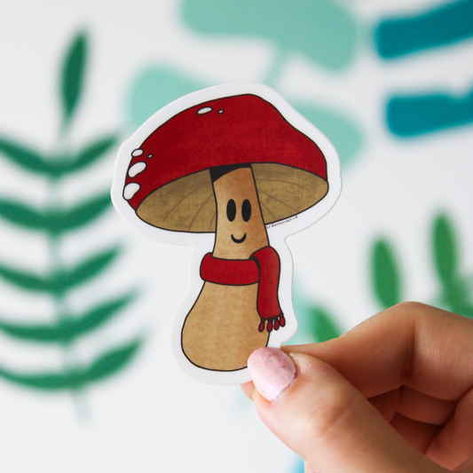 A blurred leaf pattern background. In the front is a hand holding a mushroom sticker that has a red cap and matching red scarf with a smile on its face.
