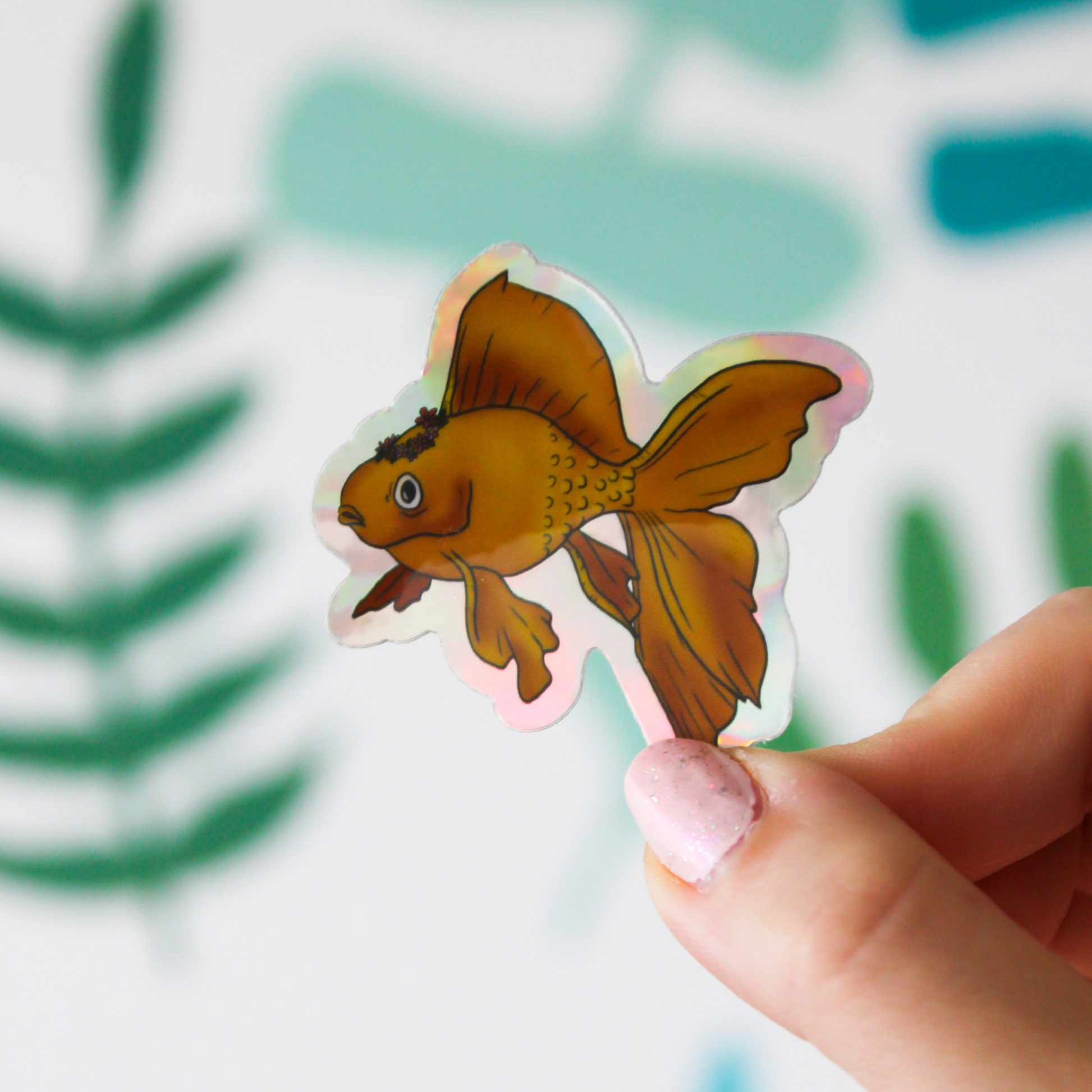 A blurred leaf pattern background. In the front is a hand holding a holographic sticker that has an orange goldfish wearing a pink flower crown.