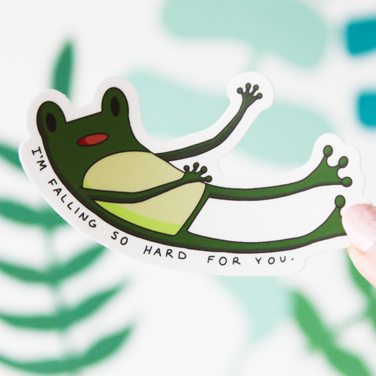 A hand holding a sticker of a green cartoon frog falling with the words "I'M FALLING SO HARD FOR YOU." underneath. A blurred leaf pattern is in the background.