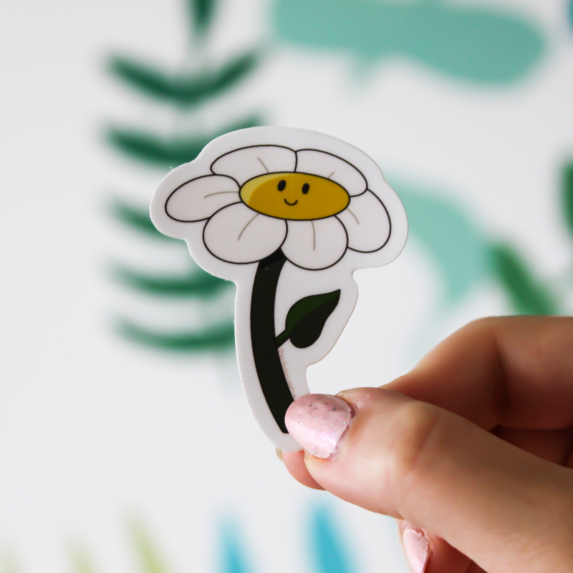 A blurred leaf background. In front is a hand holding a flower sticker with white petals, a yellow centre with a smiling face and a dark green stem.