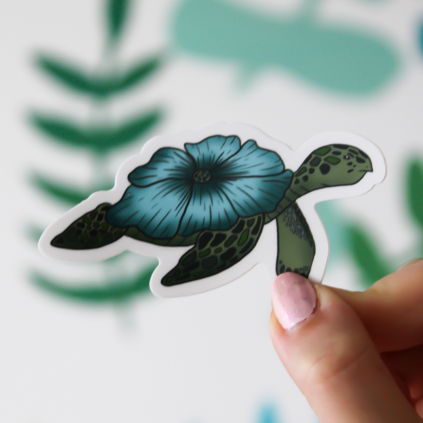A blurred leafy pattern in the background. In the front is a hand holding a turtle sticker with a blue flower on it in place of a shell.