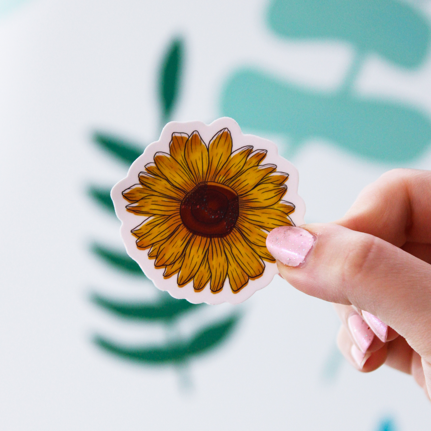 Blurred out leaf pattern in the background with a hand holding a yellow sunflower sticker in the front