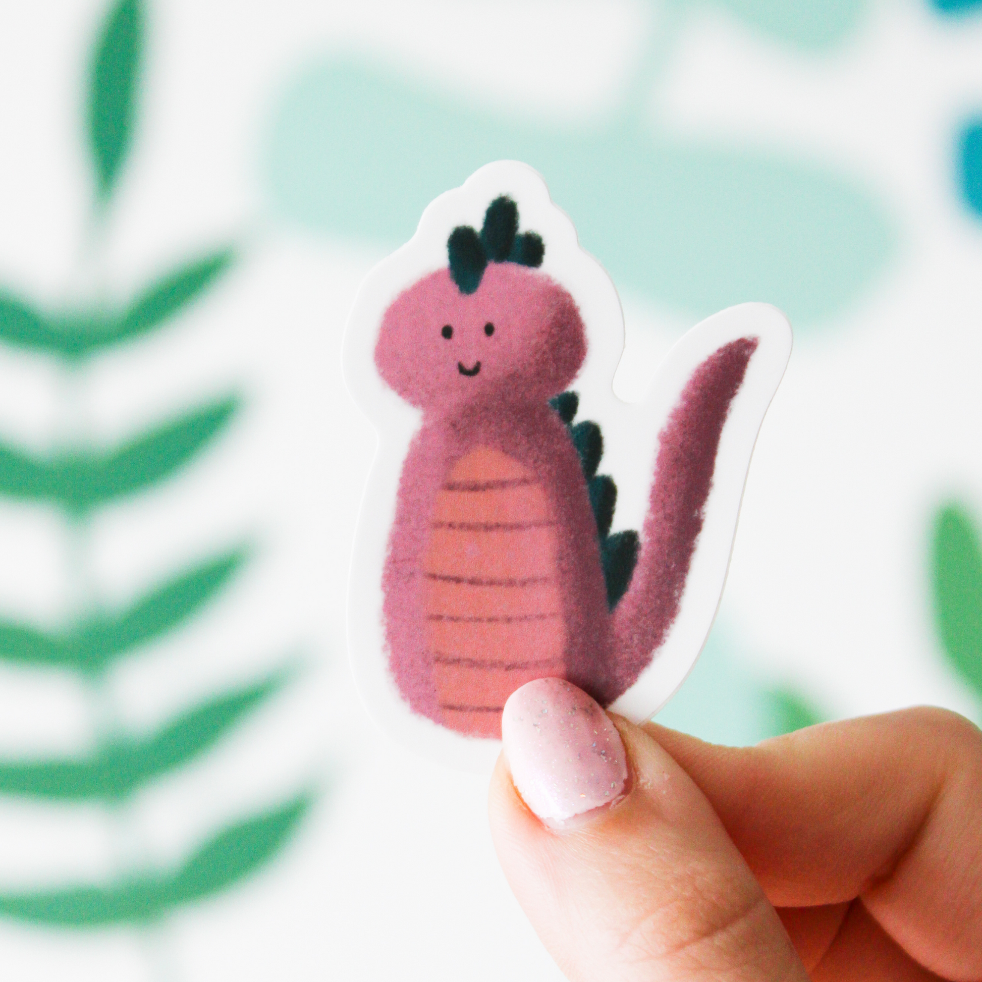 A hand holding a sticker that has a smiling pink monster  on it with dark green spikes and a long pink tail. The background is a blurred leaf pattern. 