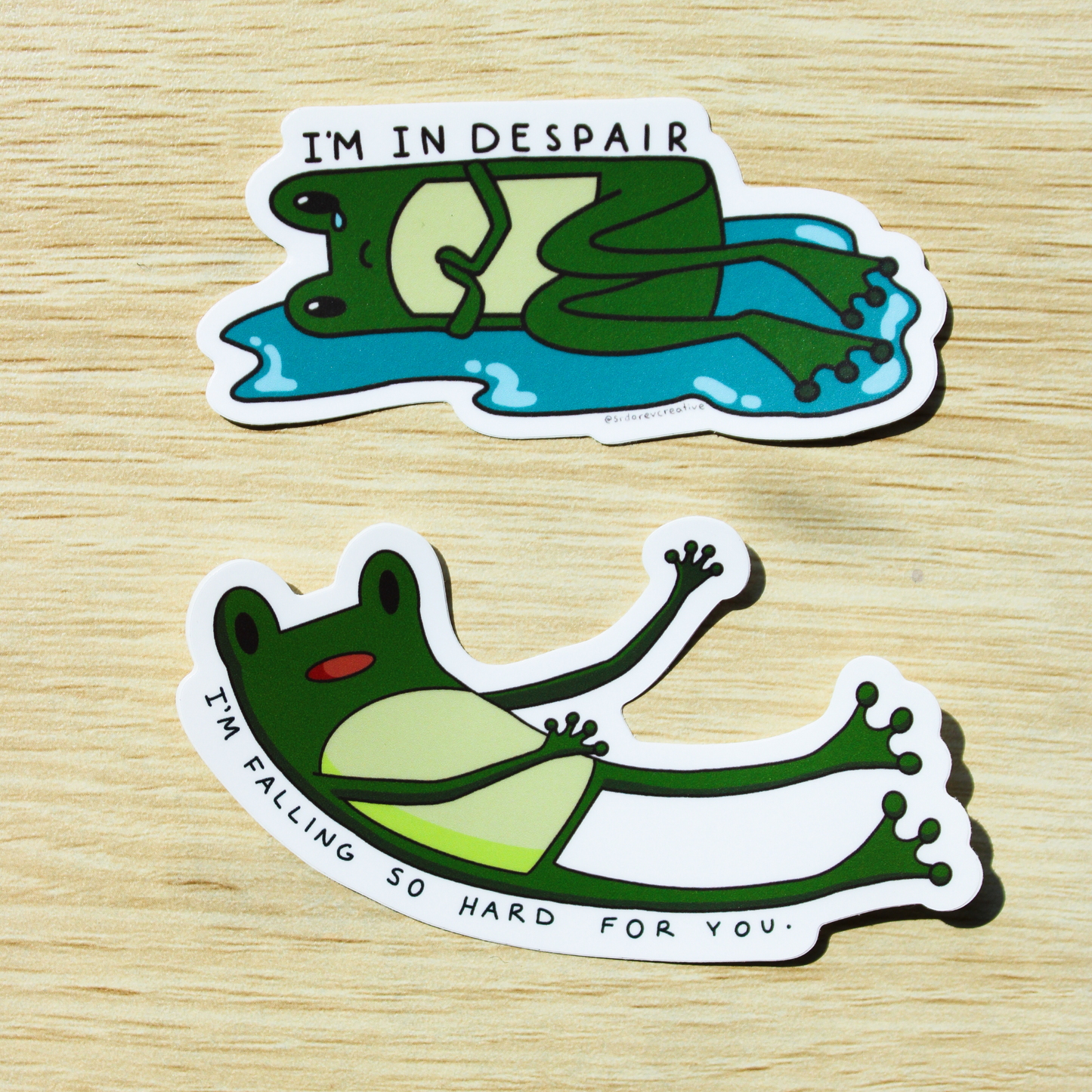 Hand holding a sticker of a cartoon frog lying down in a puddle with a tear falling down one eye and holding its hands. On top is written "I'M IN DESPAIR". On the bottom is another frog sticker of a cartoon frog falling with "I'M FALLING SO HARD FOR YOU" written underneath. Background is a wooden table.