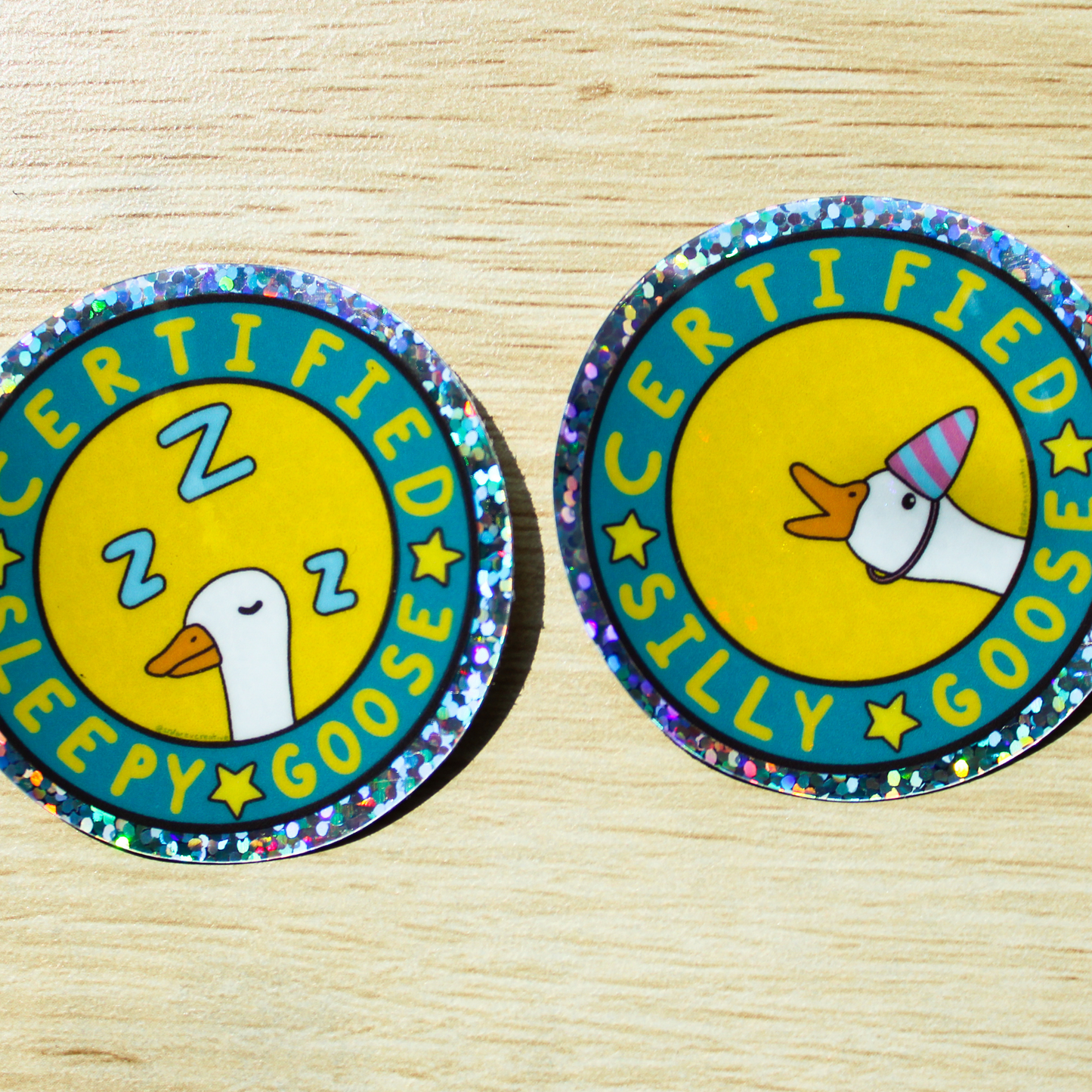 Two holographic circle glitter stickers are on a wood background. One with Certified Silly Goose Sticker written in a circle with stars in between the words. A goose with a blue and pink party hat is popping out on a yellow background. The other with Certified Sleepy Goose written in a circle with stars and a white goose with Z's sleeping on a yellow background.